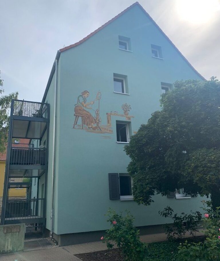 Nachher-Bild des Gebäudes 44A in der Birkenfelder Str. Die Fassade zeigt ein Gemälde: Eine Frau in historischer Kleidung spult Baumwolle auf eine Spule, während ein Kind mit einer Puppe spielt. Das Gebäude wurde umfassend renoviert und erstrahlt in einem hellen Blau-Grünton, zudem wurden Balkone hinzugefügt.