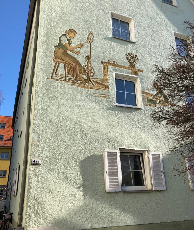 Vorher-Bild des Gebäudes 44A in der Birkenfelder Str. Die Fassade zeigt ein Gemälde: Eine Frau in historischer Kleidung wickelt Baumwolle auf eine Spule, während ein Kind mit einer Puppe spielt. Das Gebäude hat einen grünlichen Ton, der im Laufe der Zeit verblasst ist. In der untersten Etage sind Fenster mit alten Türen, bei denen die Farbe abblättert.