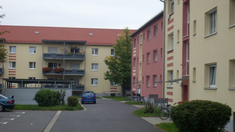 Das Nachher-Bild eines Gebäudes in der Gerhart-Hauptmann-Straße 1-11. Moderne Balkone in dunklem Grau wurden hinzugefügt, während das Gebäude in einem Pastellgelb und -rot erstrahlt.