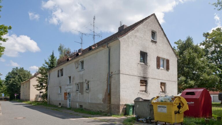 Vor der Renovierung: Gebäude in der Herderstraße 3. Ein älteres, renovierungsbedürftiges Gebäude, dessen einst weiße Fassade von Schmutz verunreinigt ist. An einigen Stellen ist der Putz abgebröckelt und Risse sind erkennbar. Einige der Fenster haben alte Türen, die quer hängen.